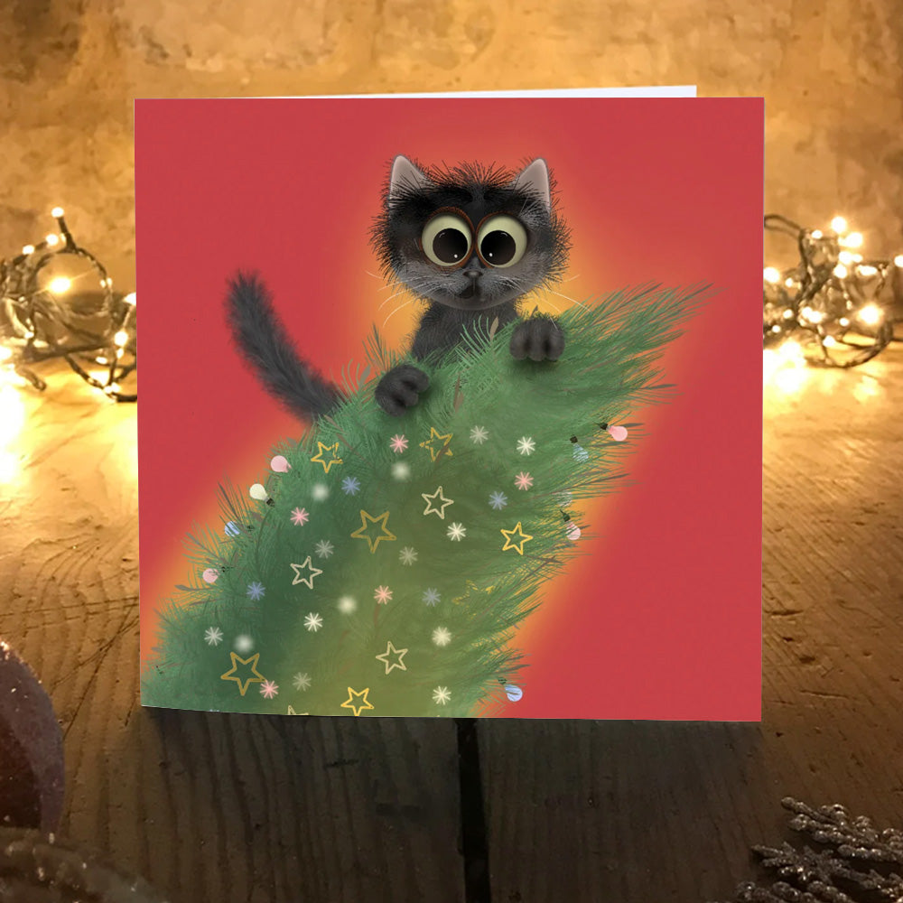Trubble Christmas card: fluffy black kitten peeking over the top of a decorated Christmas tree, on a wooden table with fairy lights